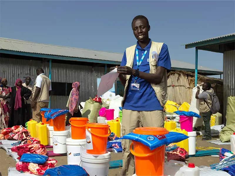 Plan International staff member standing amongst buckets, blankets, and items needed by displaced people.